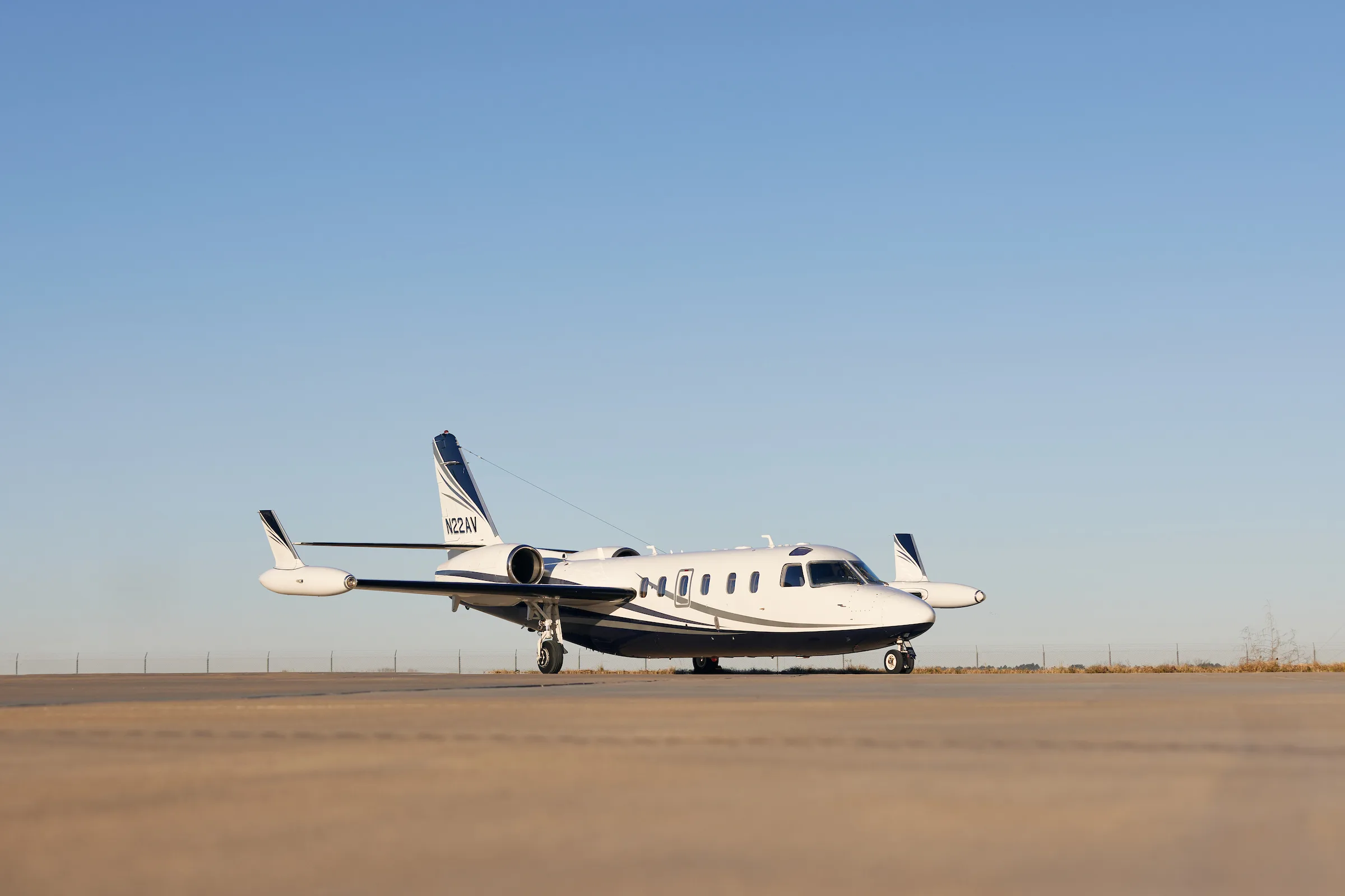 A white and blue business jet, tail number N22AV, is parked on an airfield under a clear blue sky.