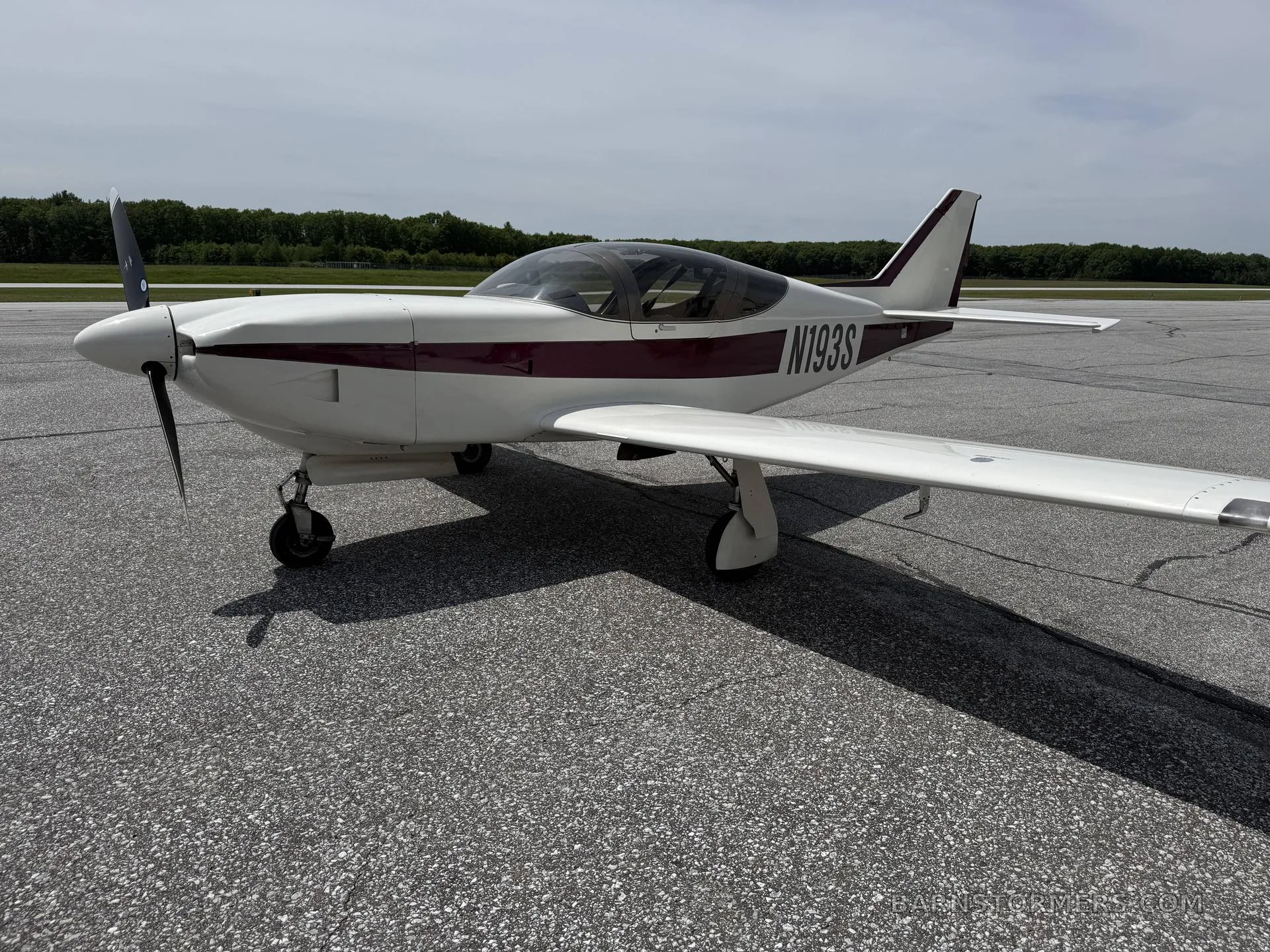 A white and maroon single-engine aircraft (N193S) with a bubble canopy is parked on an asphalt surface under a partly cloudy sky, viewed from the front-right.