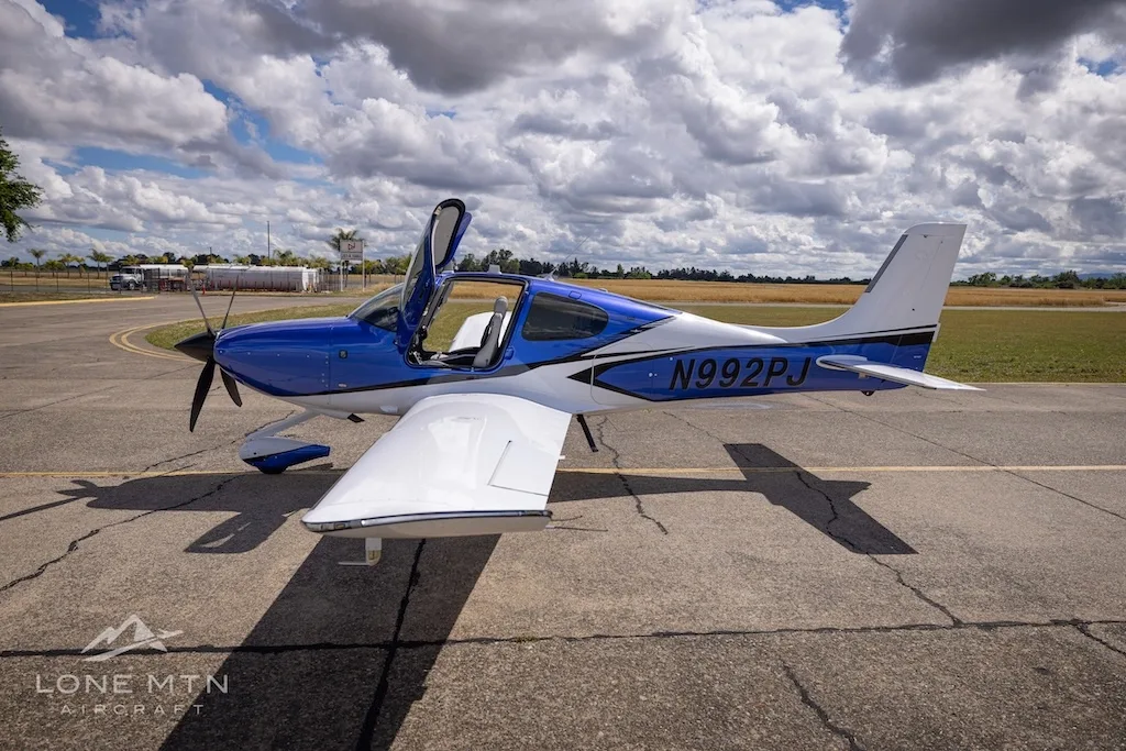A blue and white Cirrus SR22 aircraft with its door open, parked on an airfield with buildings in the background.