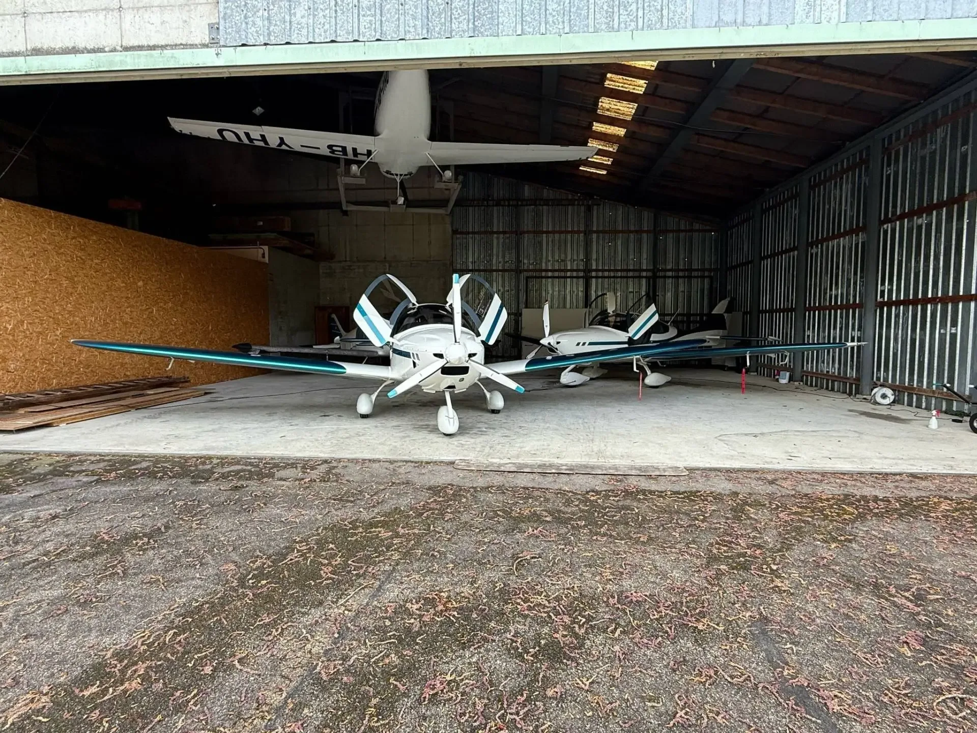 Two white and teal light aircraft are parked inside a hangar, with another white aircraft hanging from the ceiling.