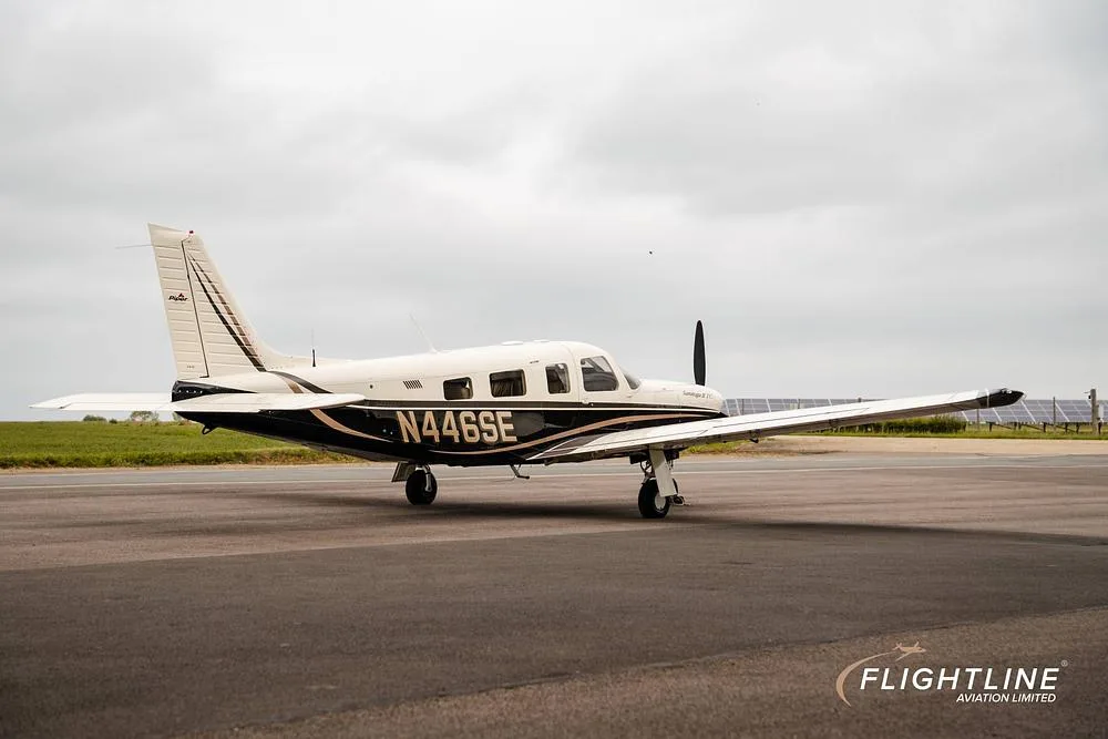 A white and black Piper Saratoga II TC, registration N446SE, parked on an asphalt runway with solar panels in the background.