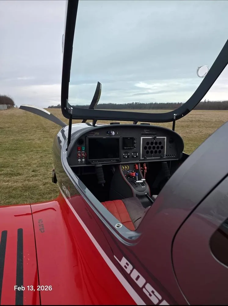 The open cockpit of a red and grey BOSSA 915iS aircraft, showing the instrument panel and controls.