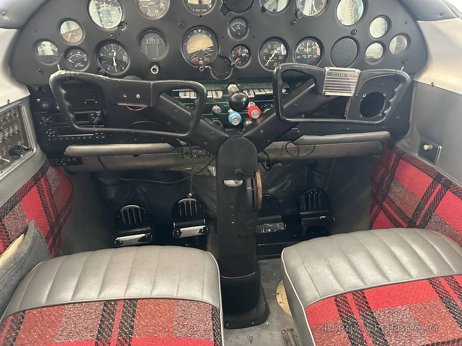 The cockpit of an aircraft, showing the instrument panel, control yokes, and front seats upholstered in gray and red plaid fabric.