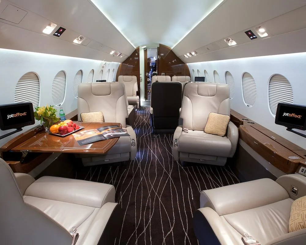 The luxurious interior cabin of a business jet, featuring beige leather seats, a wooden table with refreshments, and monitors displaying "jeteffect".