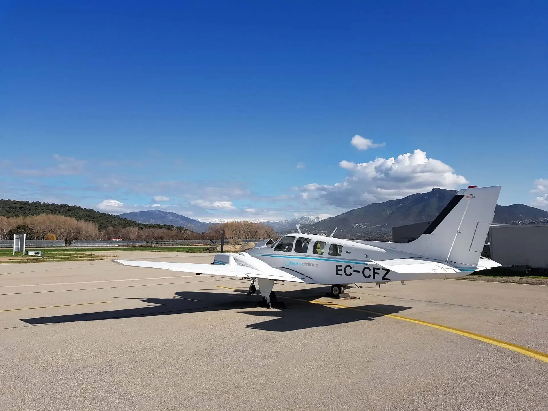 Side view of a white twin-engine propeller aircraft with blue stripes and registration EC-CFZ on an airport tarmac with mountains.