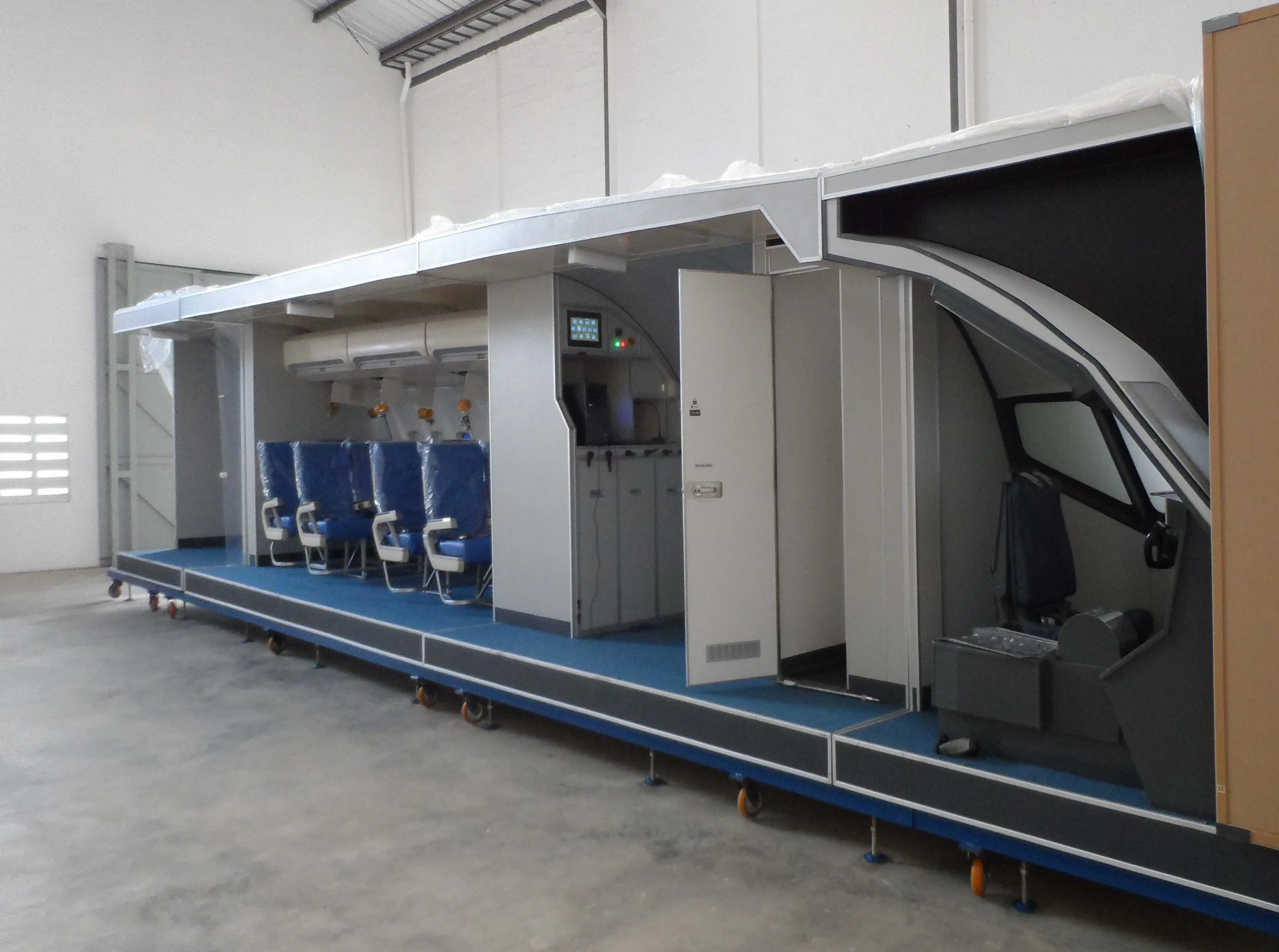 A side view of a long aircraft cabin and cockpit training mock-up with blue seats and a control panel.