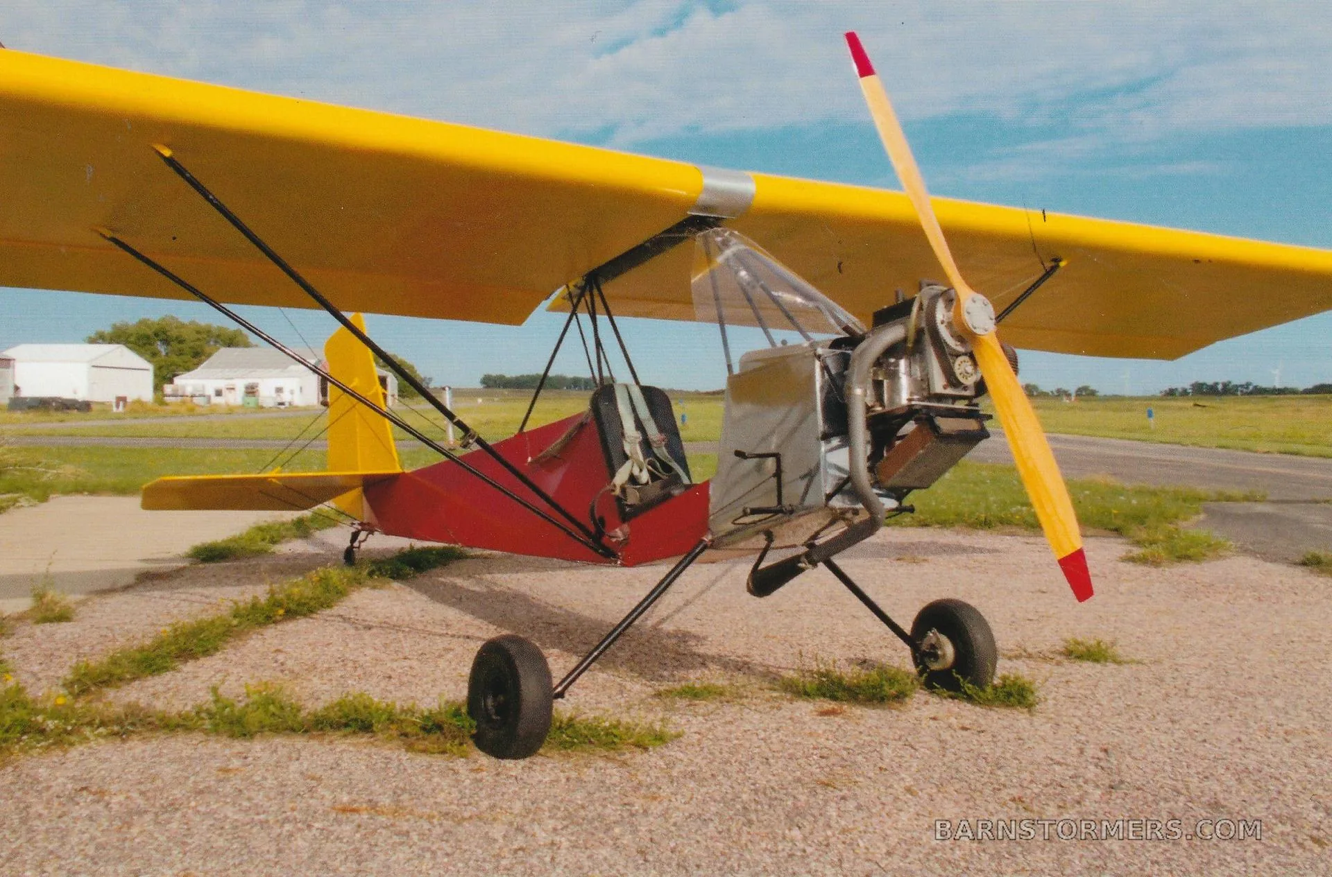 A yellow-winged, red-bodied ultralight aircraft with an exposed engine and wooden propeller, parked on a gravel runway.