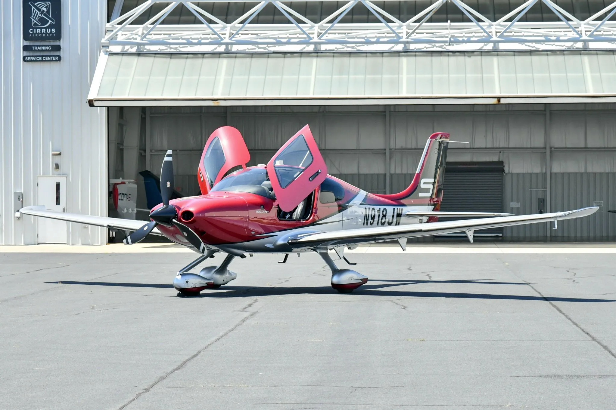 A red and white Cirrus SR22T aircraft with registration N918JW parked in front of a hangar with its doors open.