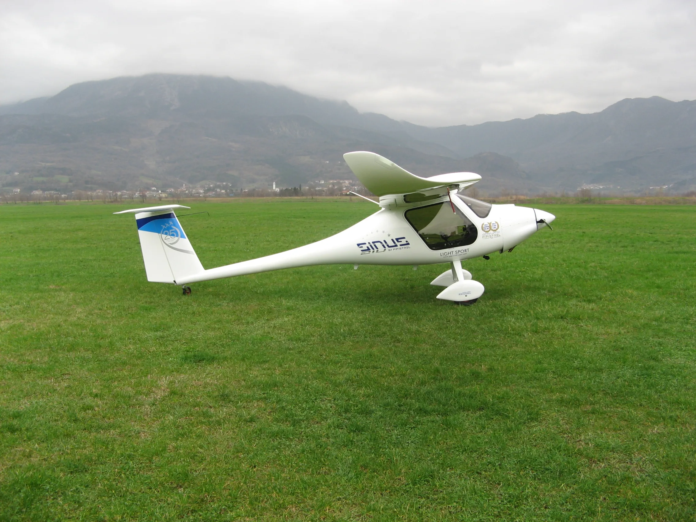 A white Pipistrel Sinus light sport aircraft with blue tail accents is parked on a grassy field, with mountains and a village in the background under a cloudy sky.