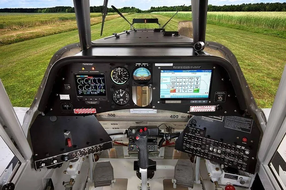 The cockpit of an agricultural aircraft, showing the instrument panel, control stick, and pedals, with a runway visible through the windshield.