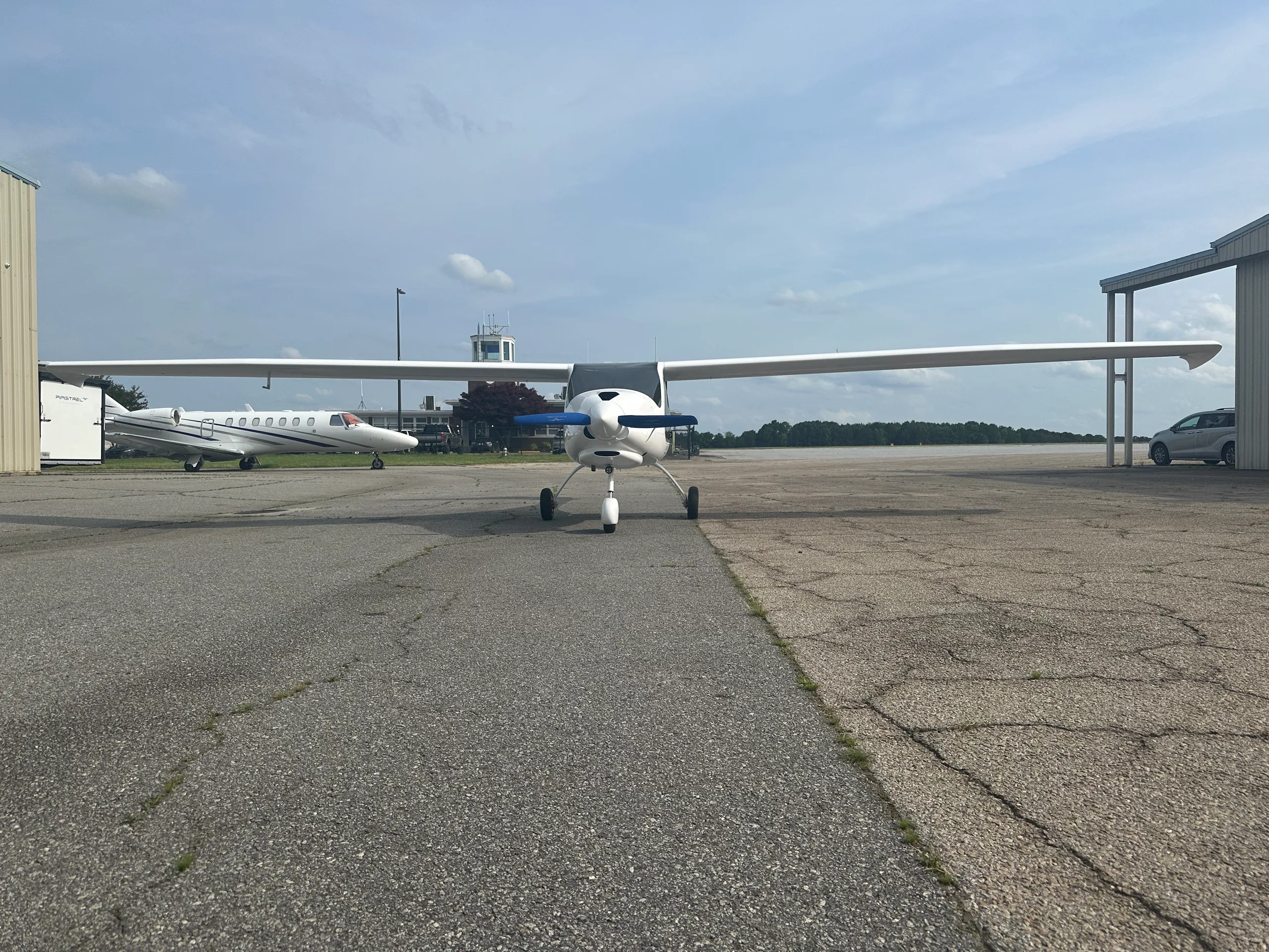 A white high-wing propeller aircraft with blue propeller tips is parked on an airport tarmac, with a jet and control tower in the background.