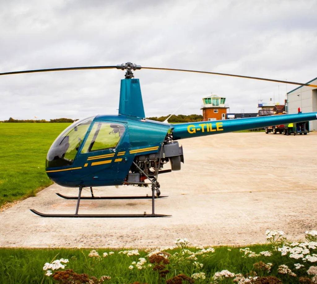 A teal Robinson R22 helicopter with yellow stripes, registration G-TILE, parked on a concrete pad at an airfield.