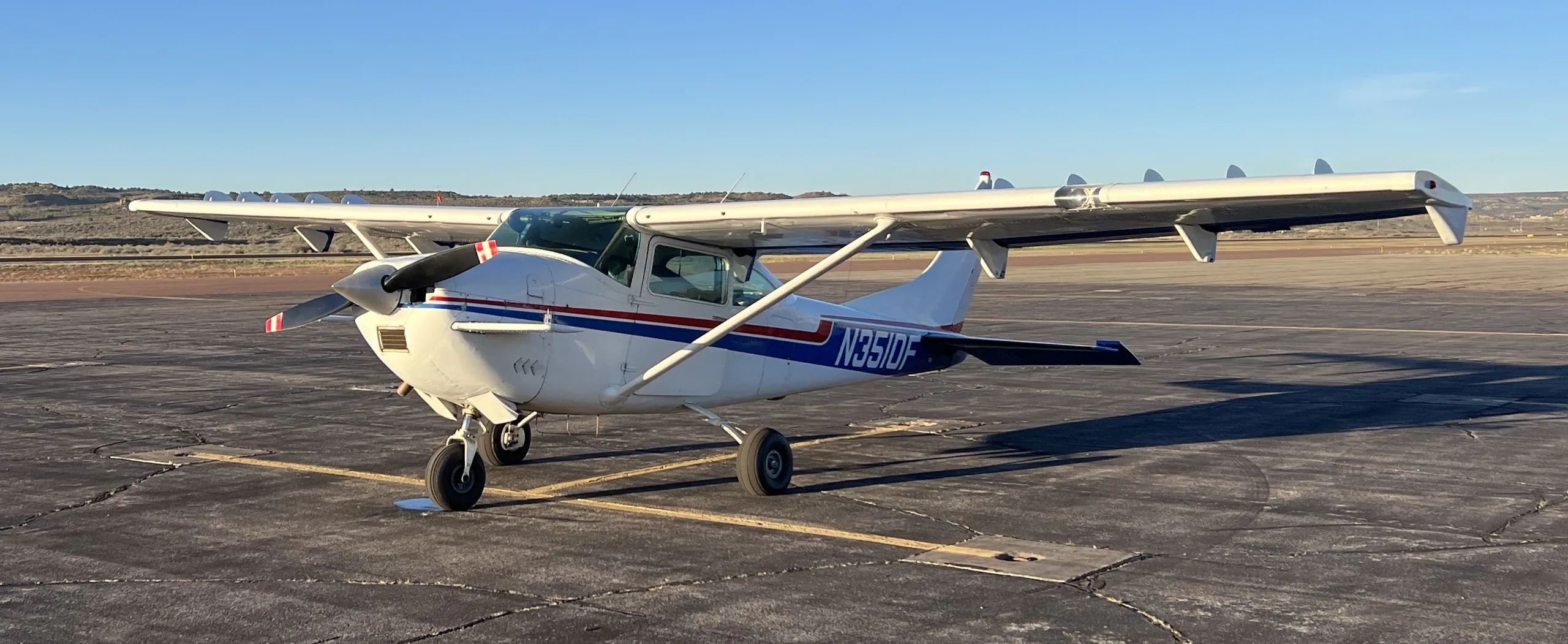 A white Cessna 182 with blue and red stripes, N3510F, parked on an asphalt apron under a clear sky.