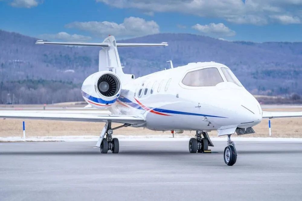 A front-quarter view of a white Learjet 45 with blue and red stripes, parked on a tarmac with mountains in the background.