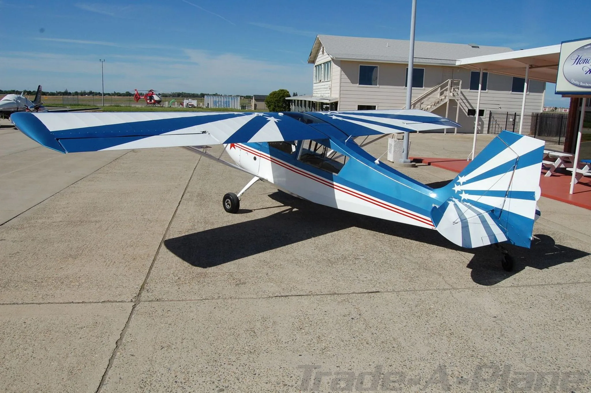 A rear-quarter view of the blue, white, and red single-engine aircraft, showing the striped tail and wings, parked on an airfield.