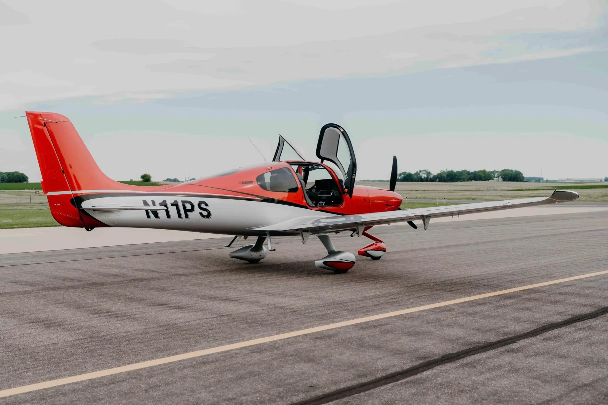 A red and silver Cirrus SR20 Premium aircraft (N11PS) with the pilot's door open, parked on an airfield tarmac.