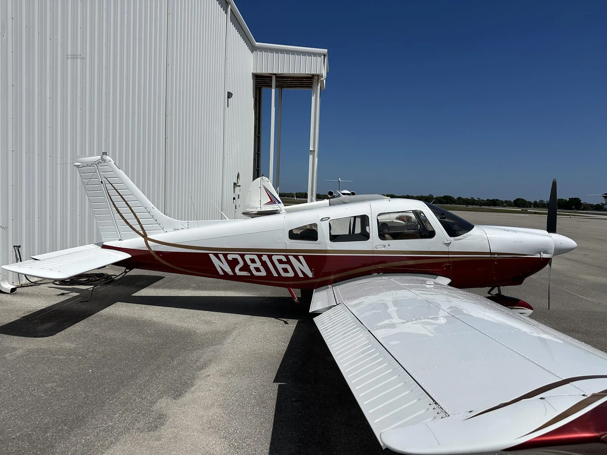A white and red Piper PA-28-161 aircraft, N2816N, parked on an asphalt ramp next to a white hangar under a clear blue sky.