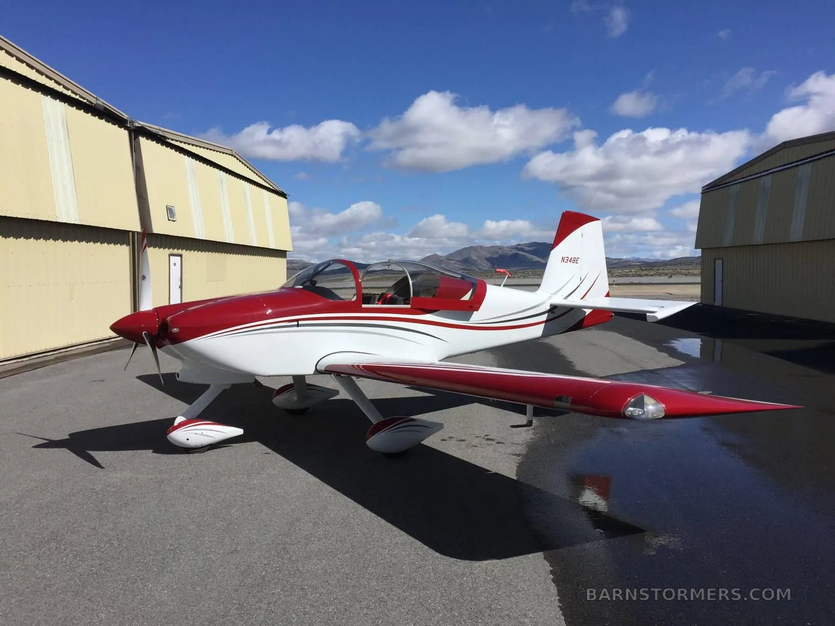 A red and white single-engine aircraft (N34BE) is parked on an asphalt ramp between two yellow hangars under a partly cloudy sky.
