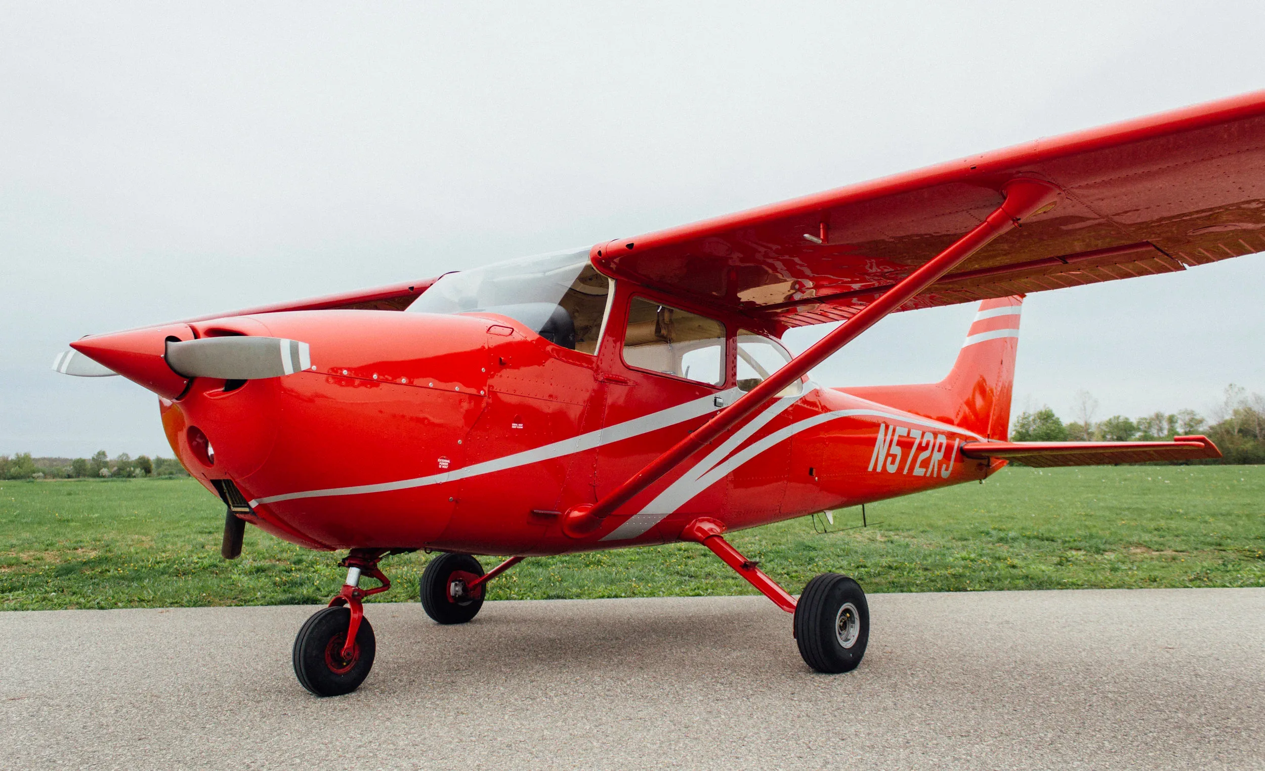 A red single-engine propeller aircraft with silver stripes, registration N572RJ, parked on an asphalt surface next to a green field under an overcast sky.