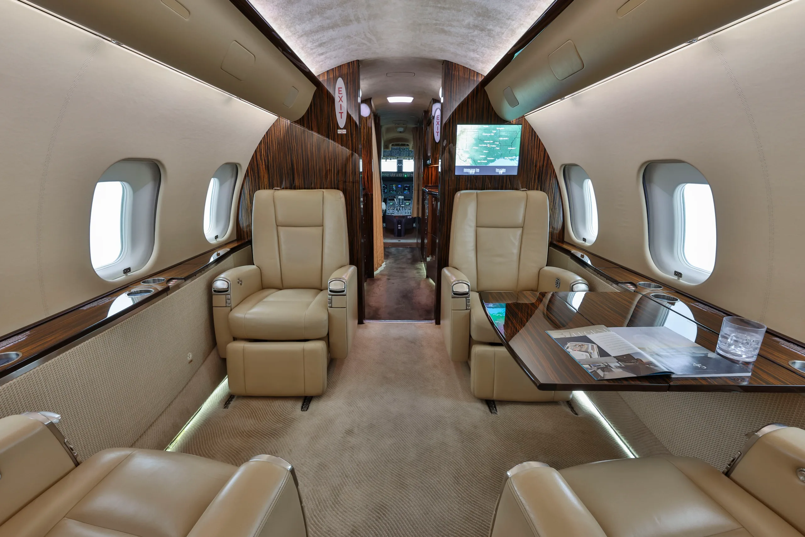 A view of the business jet's cabin, showing beige leather seating, a wooden table, and a screen displaying a map.
