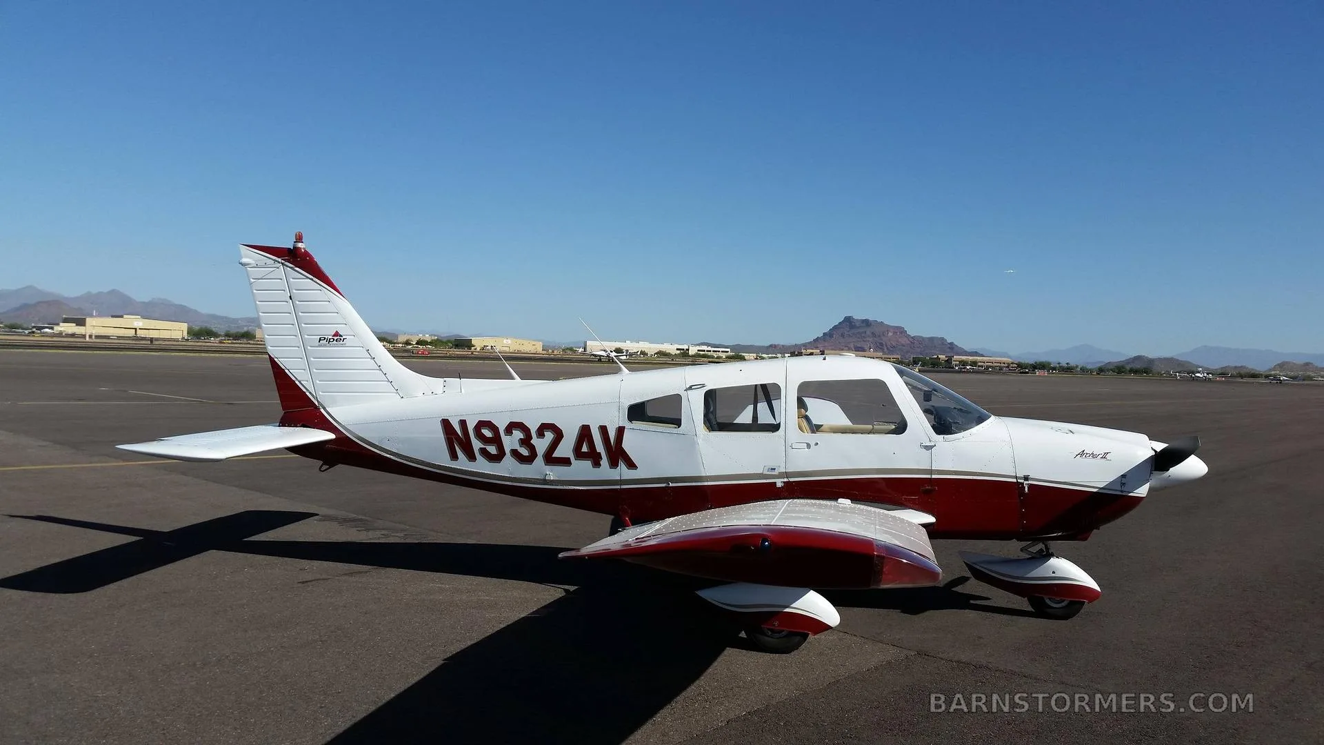 A white and red Piper Archer II (N9324K) parked on an airport tarmac under a clear blue sky.