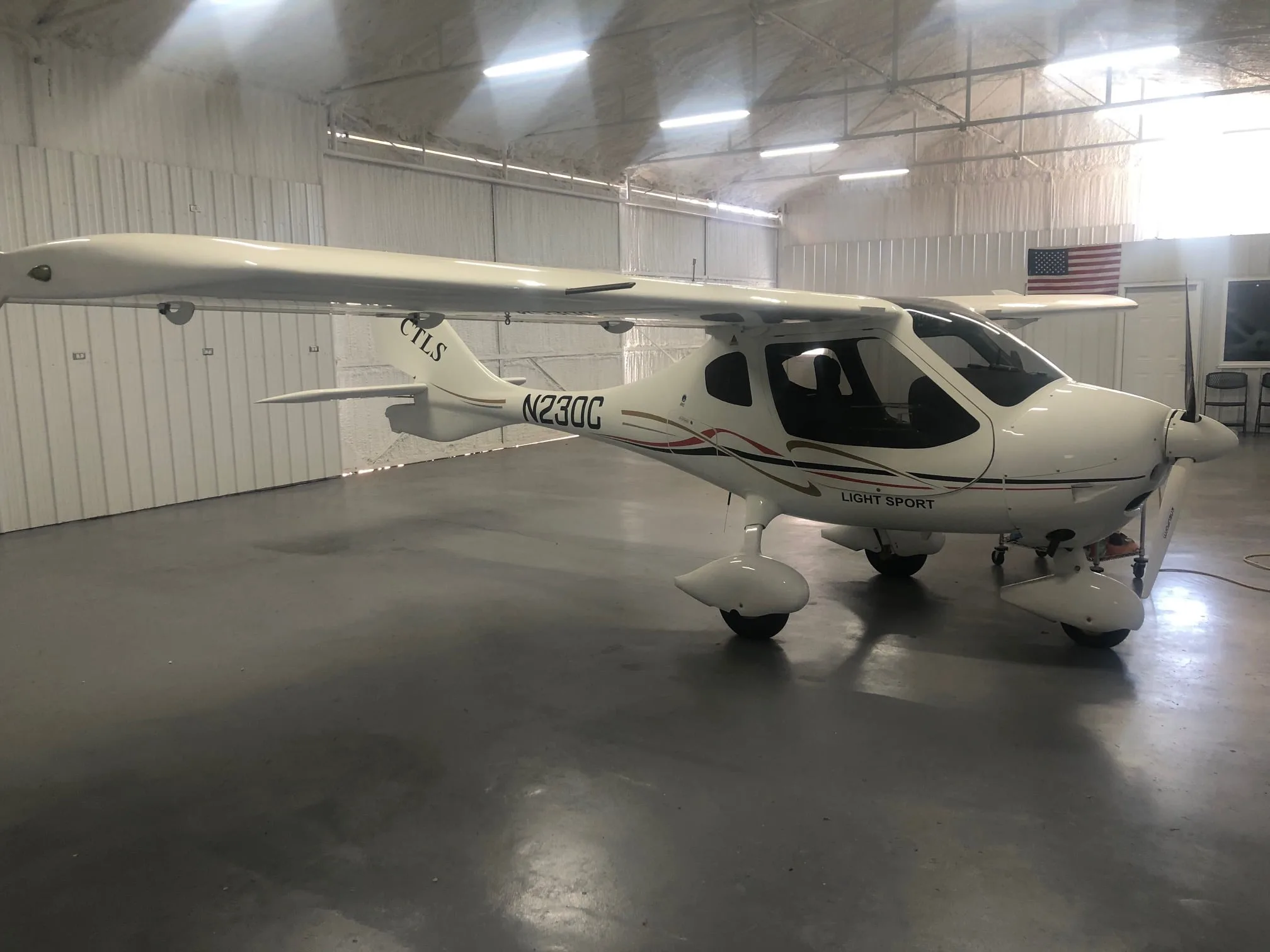 A white Flight Design CTLS light sport aircraft with red, black, and gold stripes, parked inside a hangar.