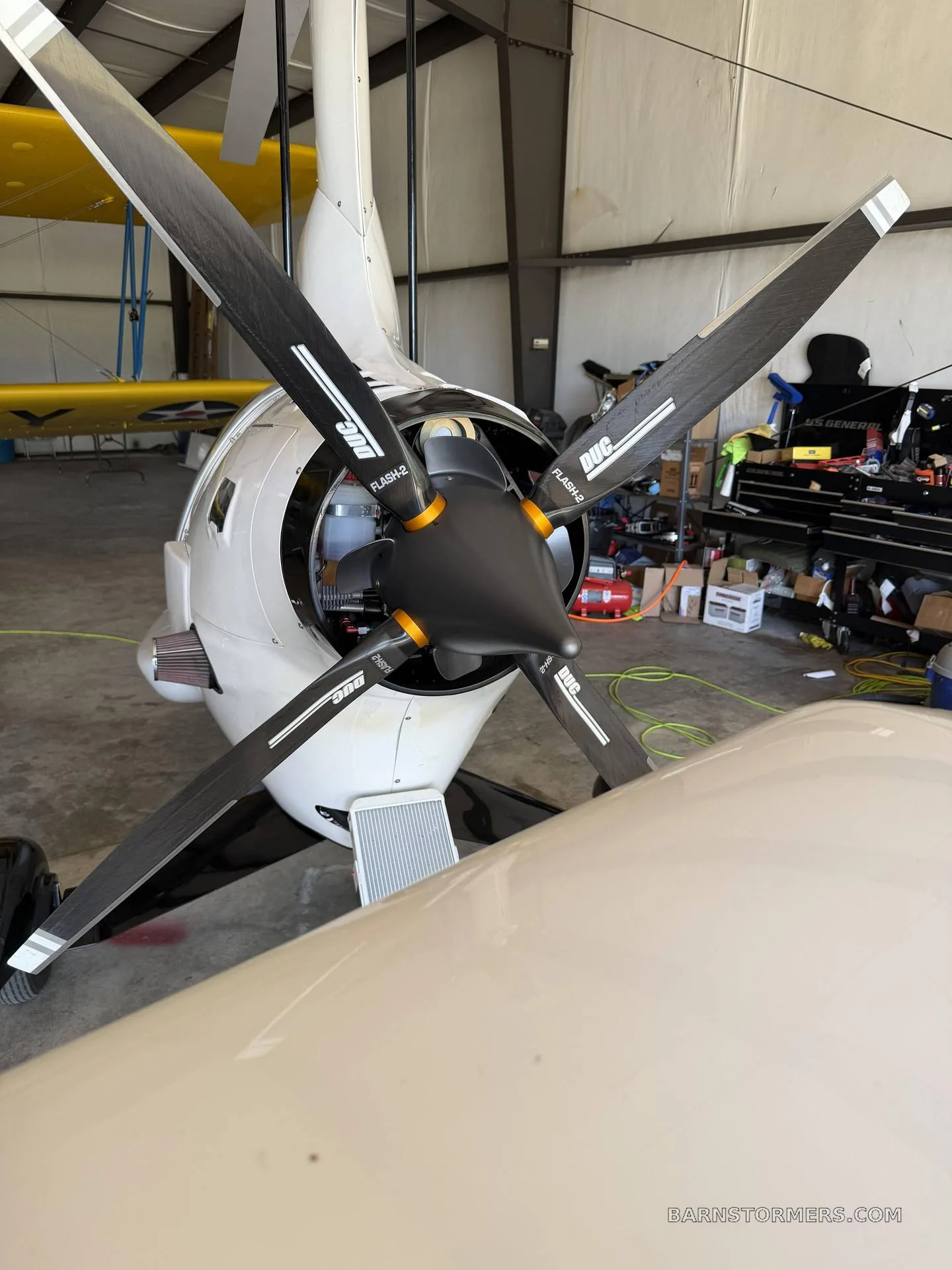 A close-up view of the four-bladed propeller and engine cowling of a white autogyro inside a hangar.