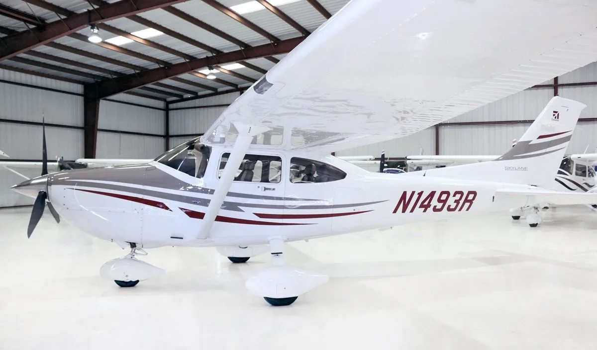A white Cessna Skylane N1493R with grey and maroon stripes is parked inside a hangar, viewed from the side.