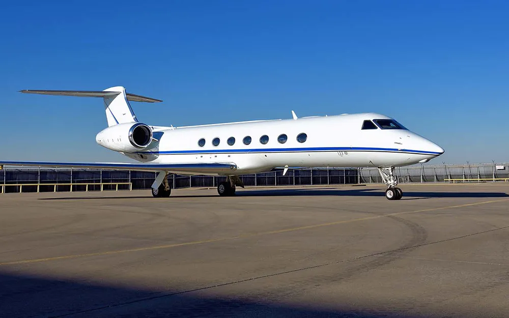 A white Gulfstream G550 business jet with a blue stripe parked on the tarmac under a clear blue sky.