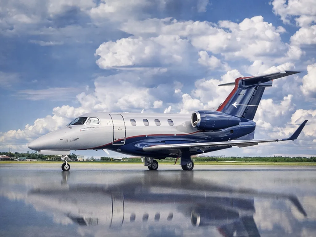 A silver and blue business jet is parked on a wet tarmac, reflecting under a cloudy sky.