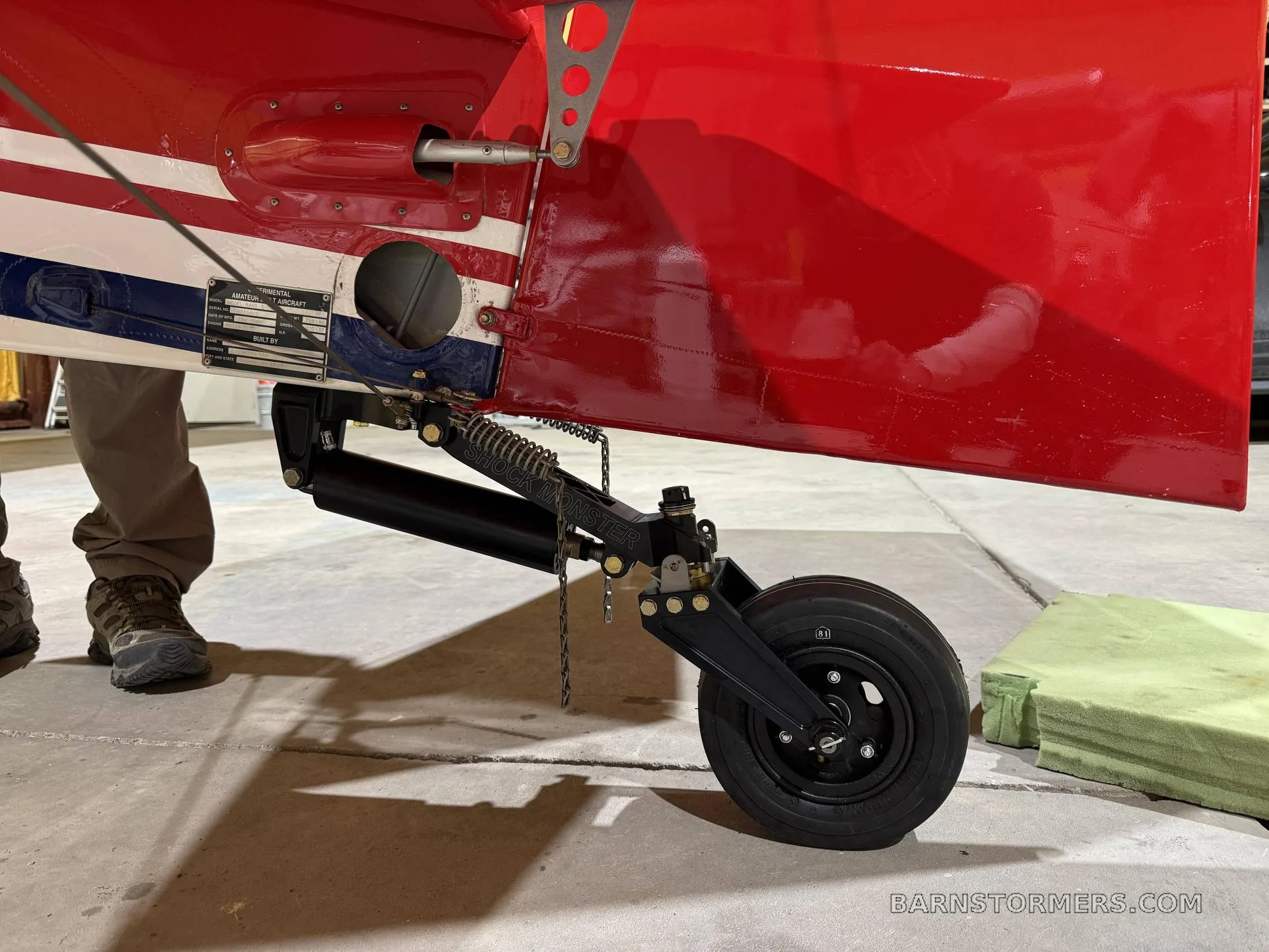 A close-up view of the black tail wheel assembly and red fuselage of a light aircraft, with a person's feet visible.
