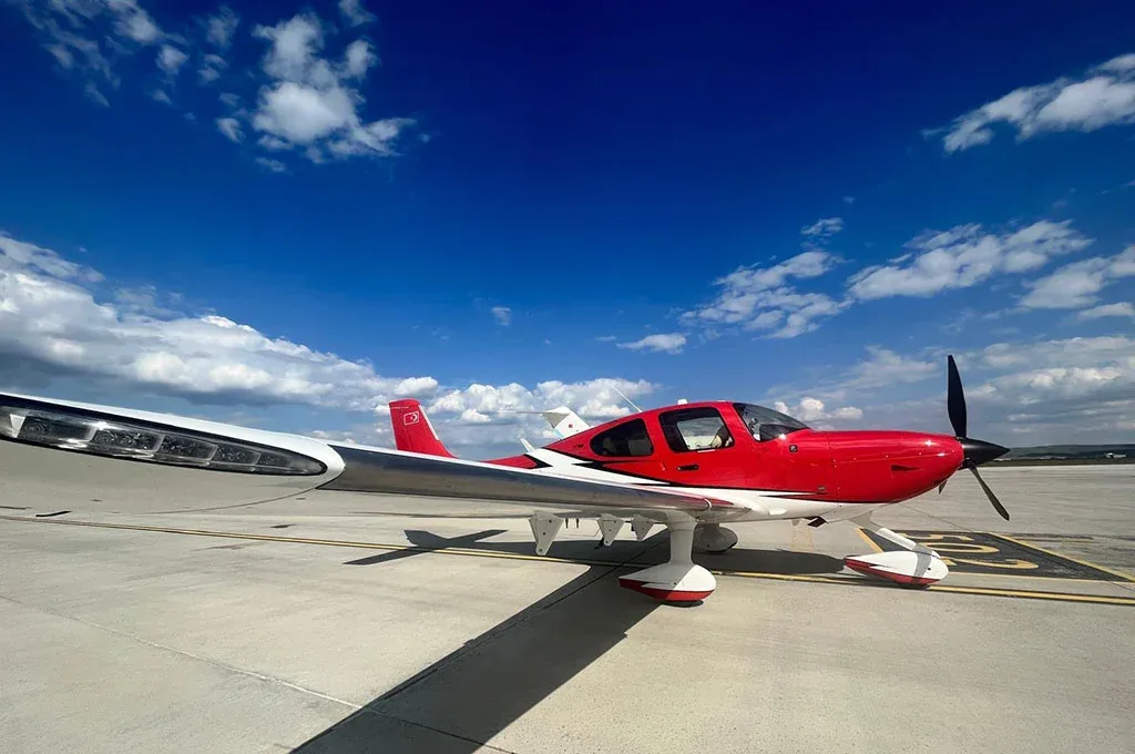 A side view of a red and white single-engine aircraft (TC-SFA) parked on an airfield under a partly cloudy blue sky.