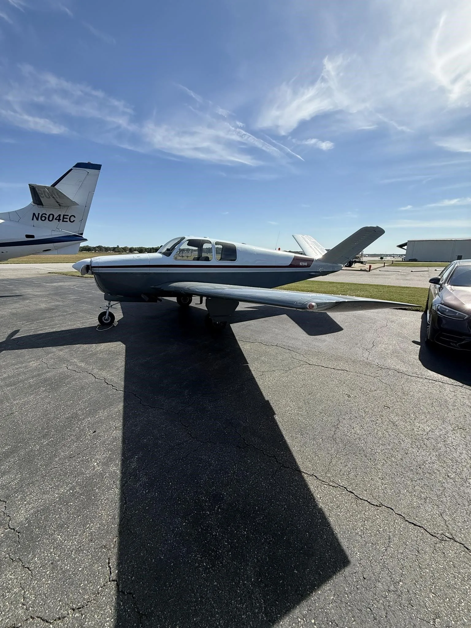 Side view of the N2194D Beechcraft Bonanza V-tail aircraft, with a white and grey paint scheme and red stripe, parked on an airfield next to another aircraft.