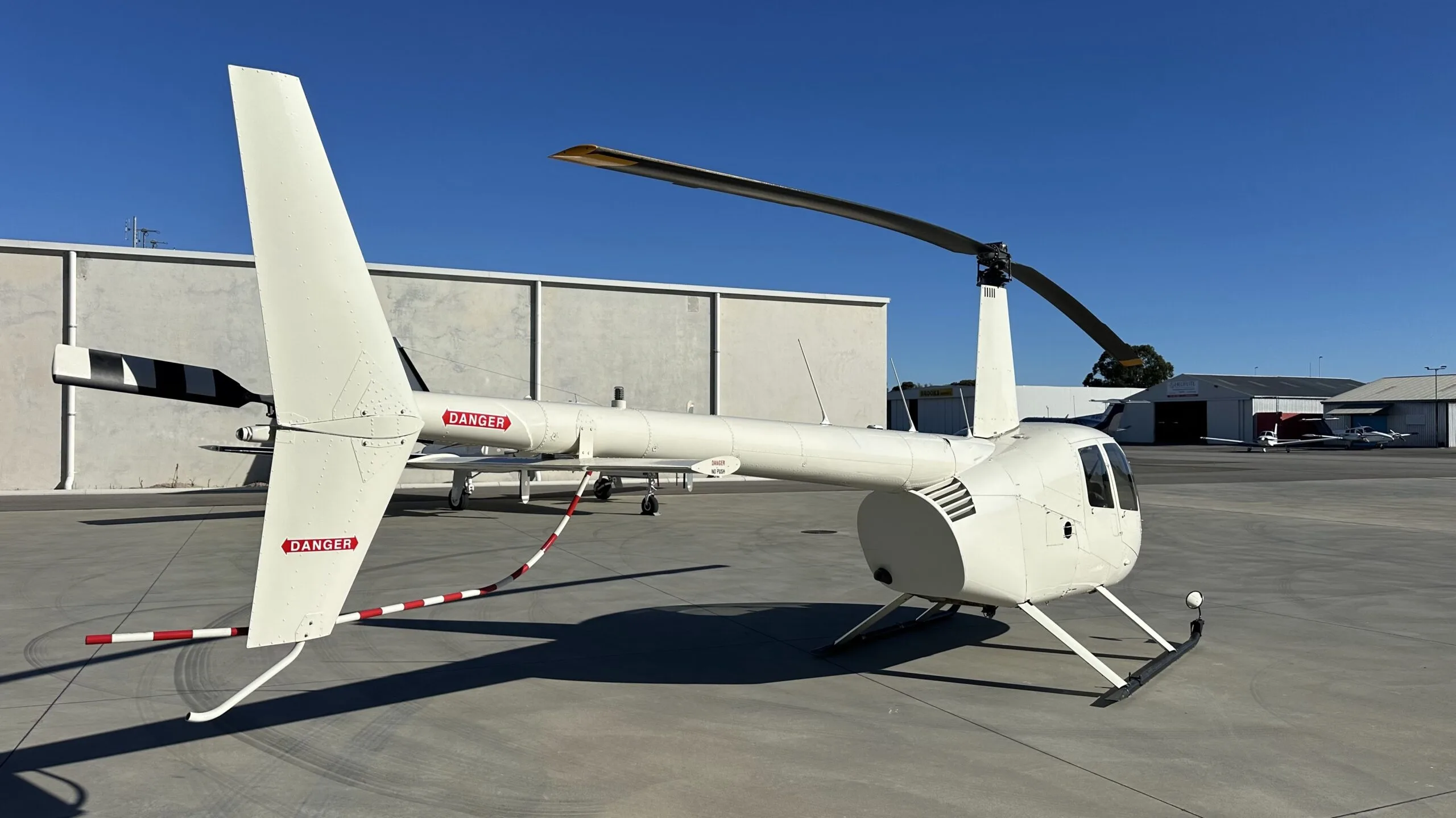A white Robinson R44 helicopter parked on an airfield tarmac, viewed from the rear-right.