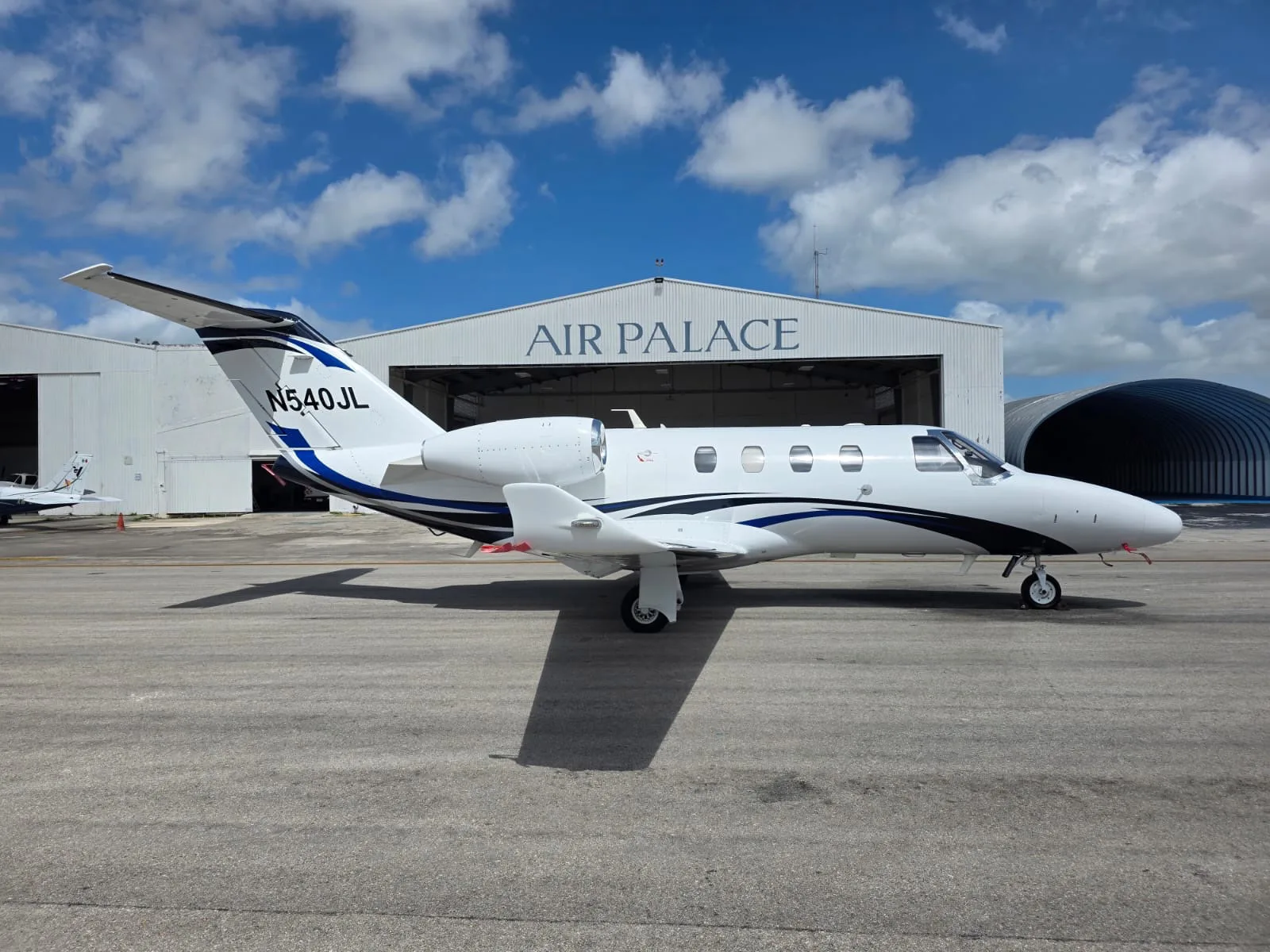 A white business jet with blue and black stripes, registration N540JL, parked on an airfield in front of a hangar under a partly cloudy sky.
