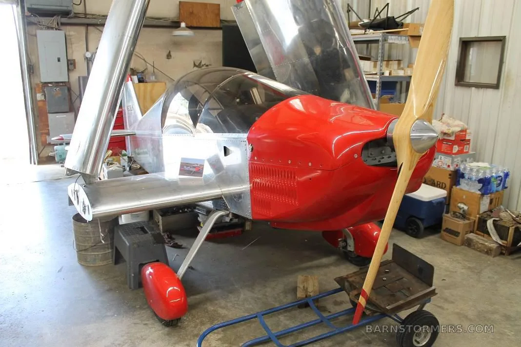 A silver and red experimental aircraft with a wooden propeller and open wings is parked in a workshop.