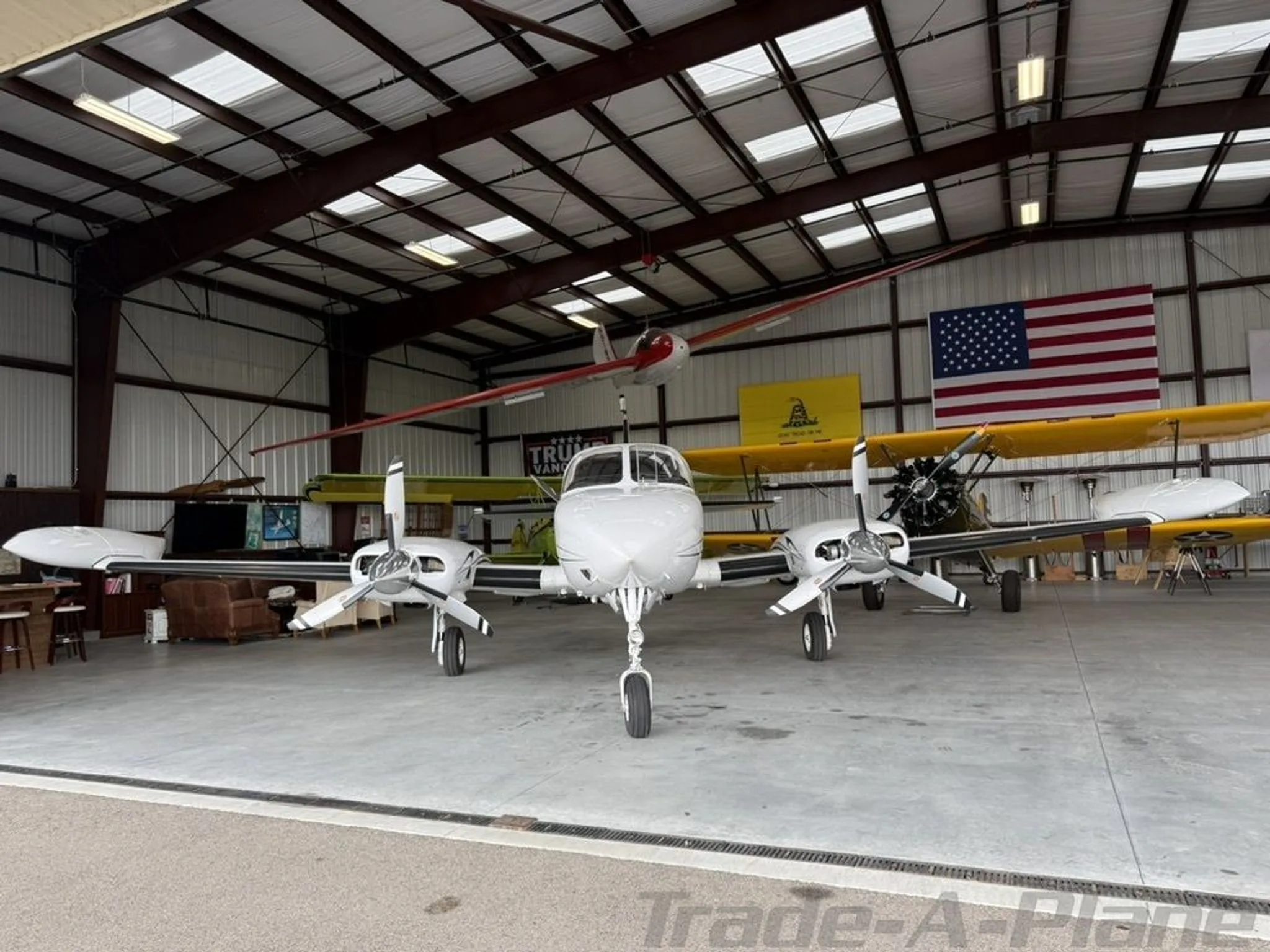 A wide front view of a white twin-engine aircraft, N653JR, parked inside a hangar with a yellow biplane and an American flag.