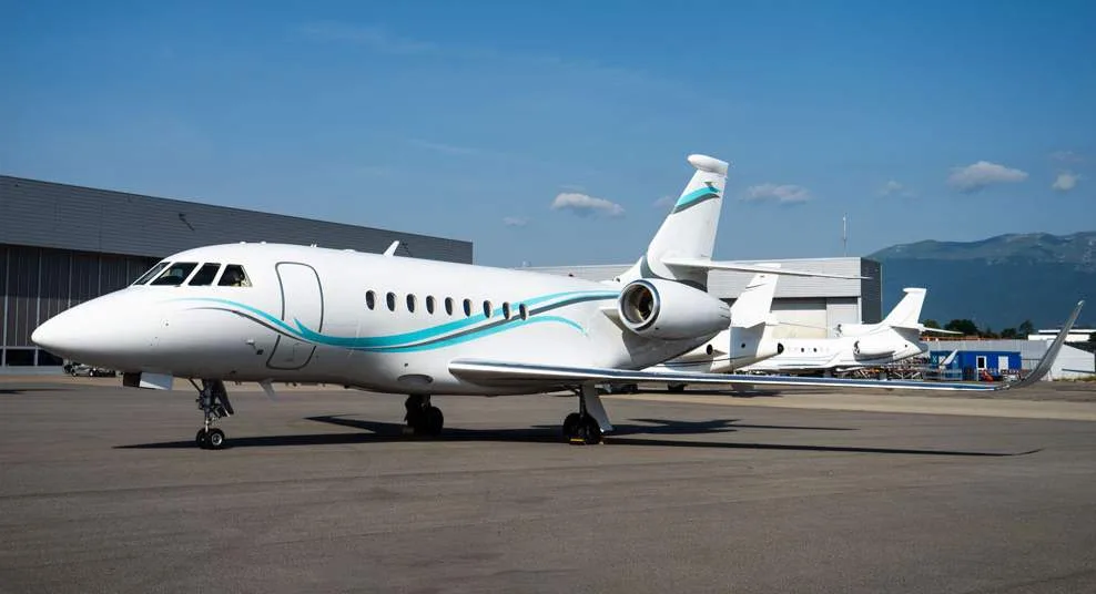 A white business jet with blue and teal stripes, parked on an airfield with hangars and mountains in the background.