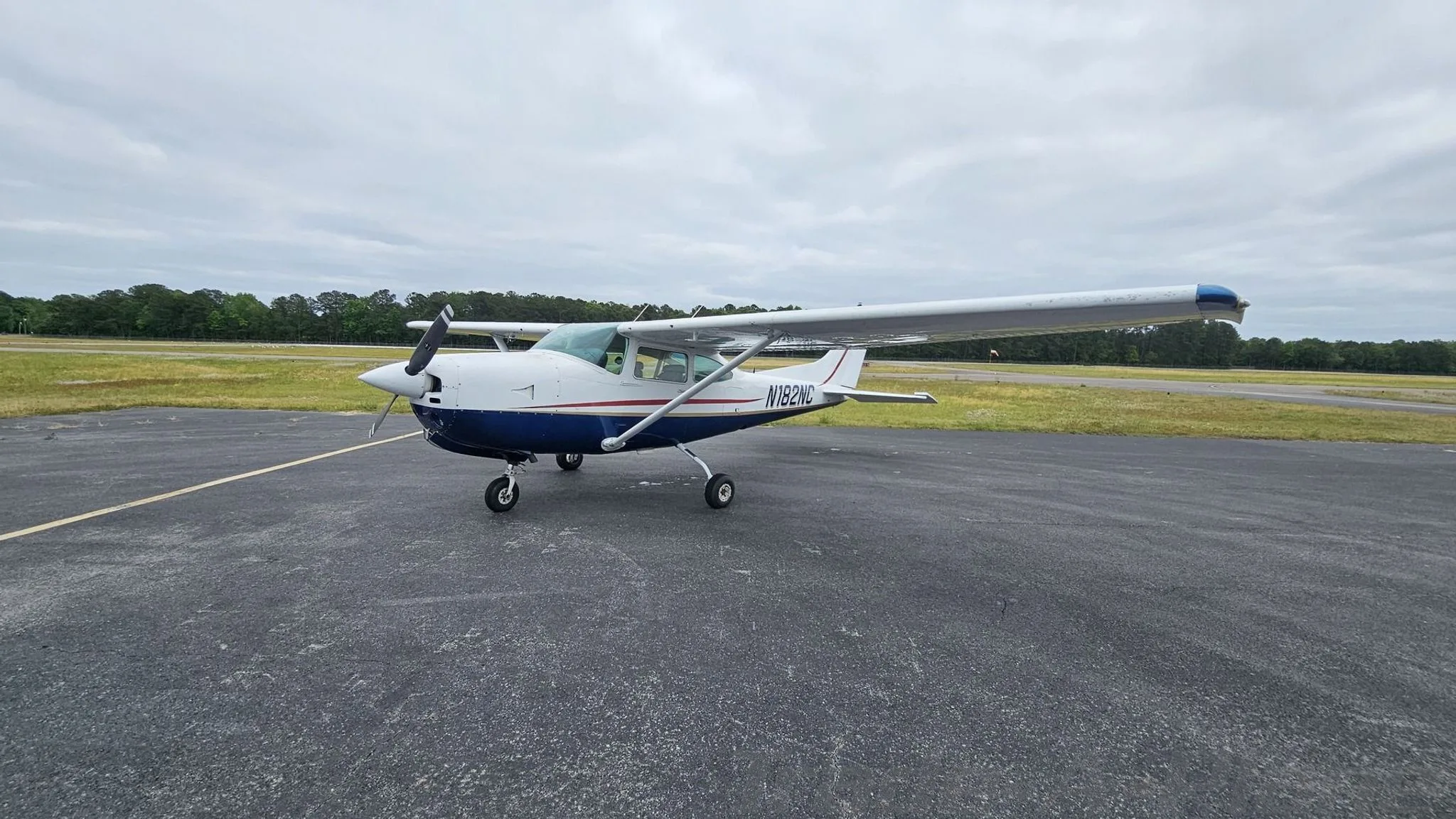 A full view of the white and blue Cessna 182 with red and gold stripes parked on an asphalt tarmac.