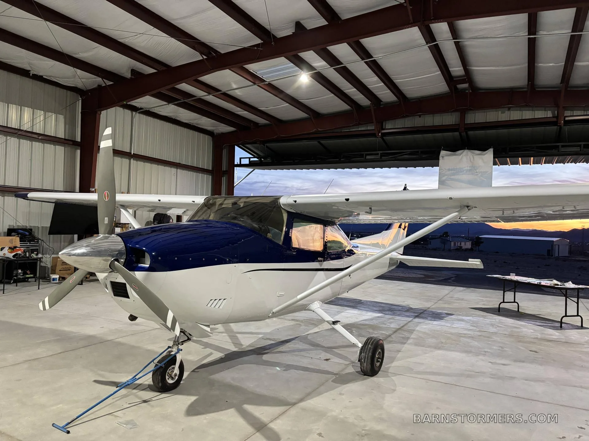 A front-quarter view of a blue and white single-engine propeller aircraft parked inside a hangar at sunset.
