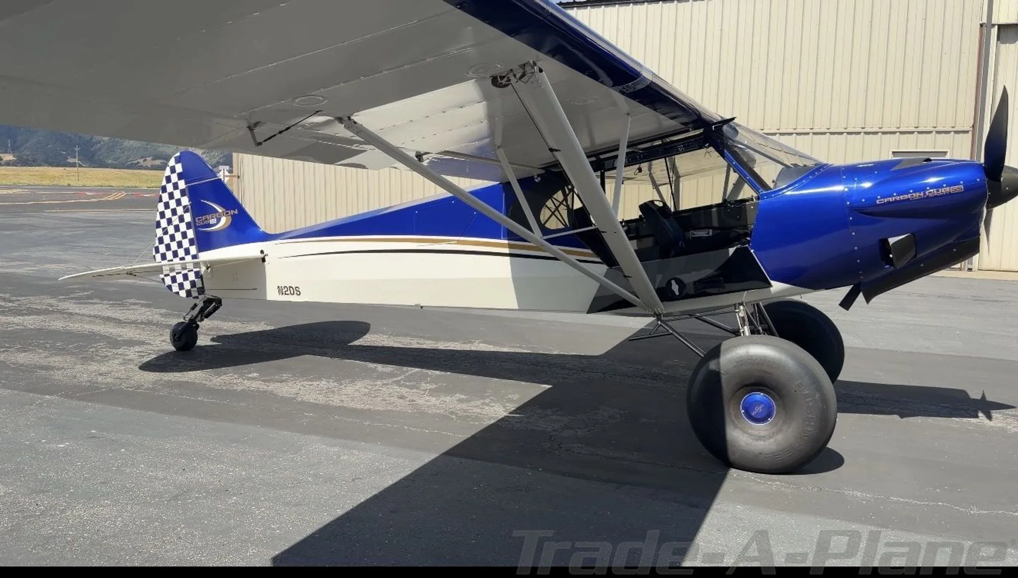A blue and white Carbon Cub SS aircraft with large tundra tires, seen from the front-right side on an airport tarmac.