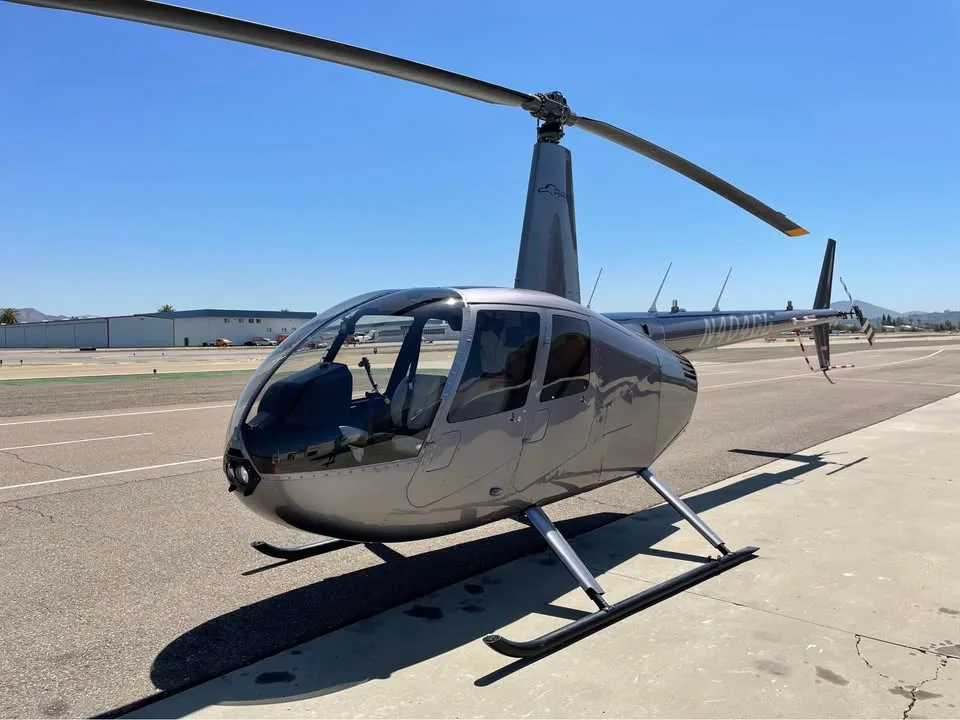 A front-right view of a grey Robinson R44 helicopter parked on an airport tarmac on a sunny day.