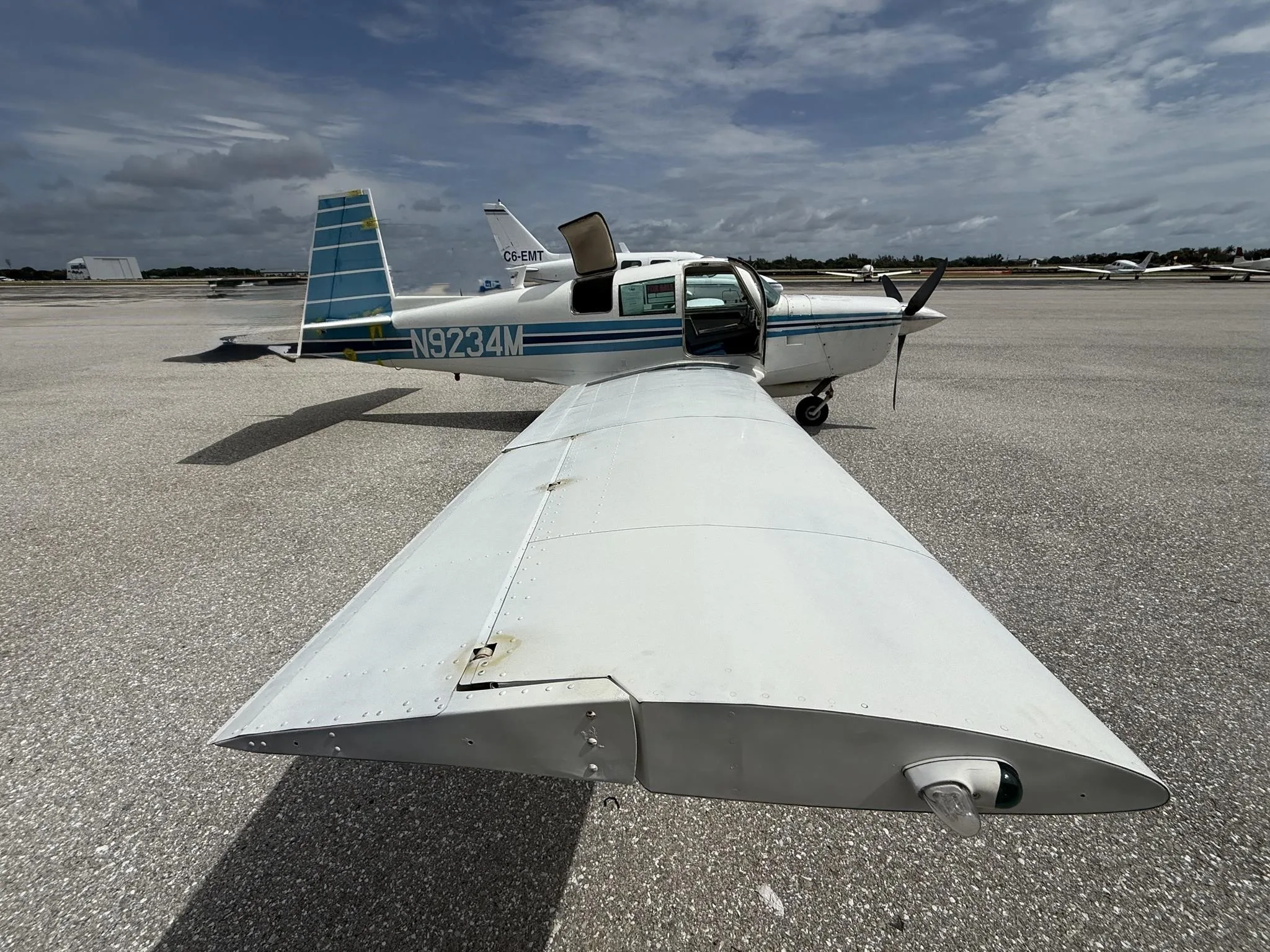 A white single-engine aircraft with blue stripes, registration N9234M, parked on an airfield with its door open under a partly cloudy sky.