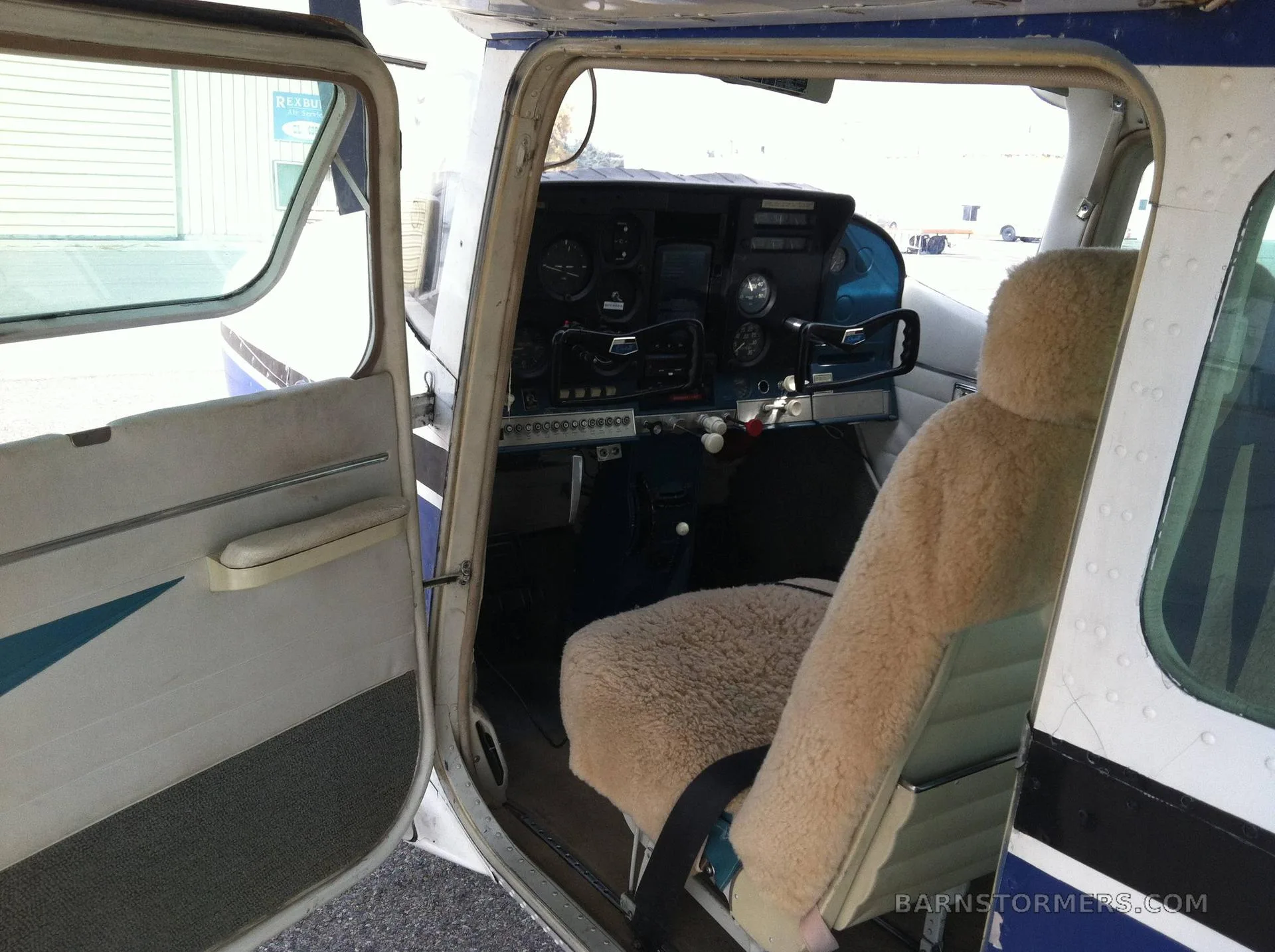 The interior of a Cessna 182 Skylane, showing a sheepskin-covered pilot's seat and part of the instrument panel through an open door.