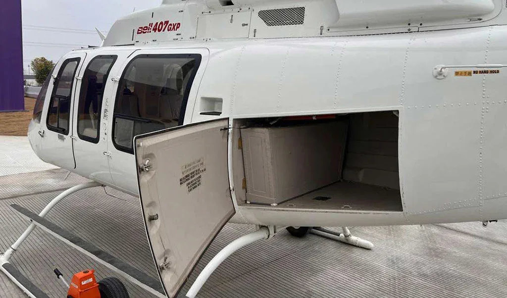 A close-up side view of a white Bell 407GXP helicopter with its cargo compartment door open, revealing an empty storage space.