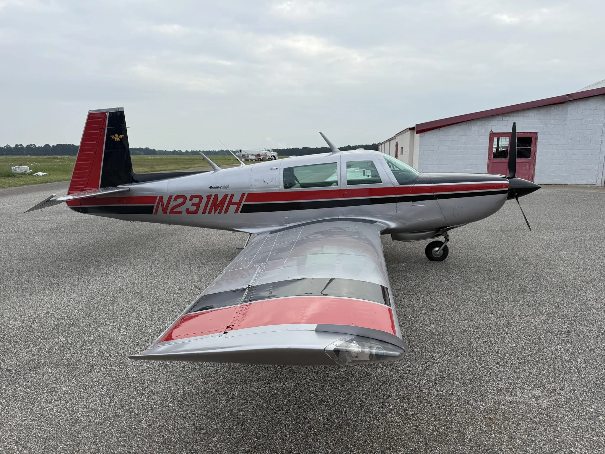 A silver Mooney 231 aircraft with red and black stripes, registration N231MH, parked on an airfield next to a white hangar.