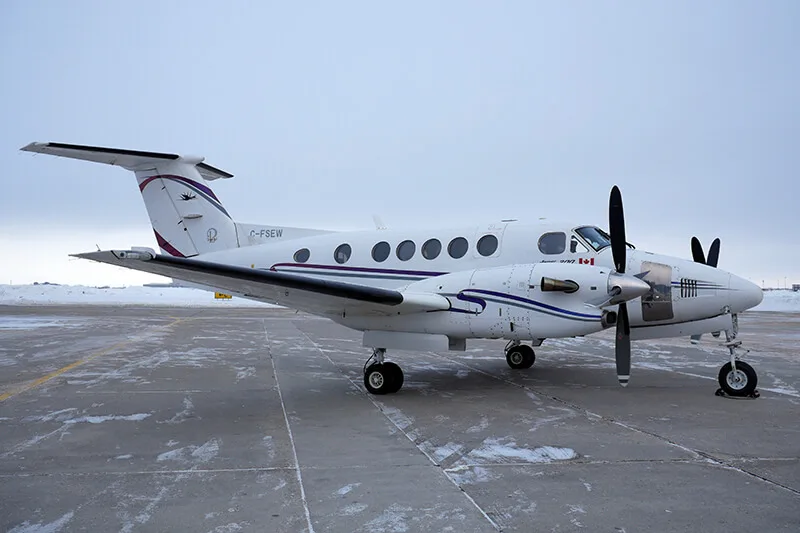 A white Beechcraft King Air 350 with purple and black stripes is parked on a snowy tarmac.
