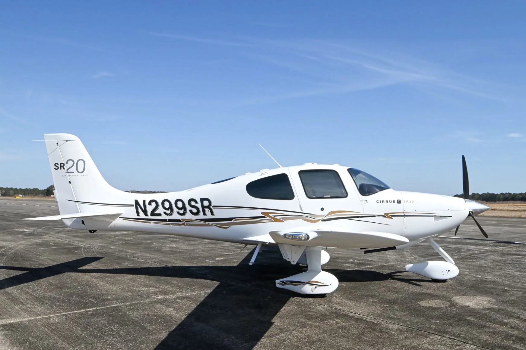 A white Cirrus SR20 aircraft with black and gold accent stripes, registration N299SR, parked on an airfield under a blue sky.