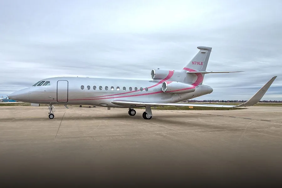 A silver Dassault Falcon 900EX private jet with pink stripes parked on an airfield.