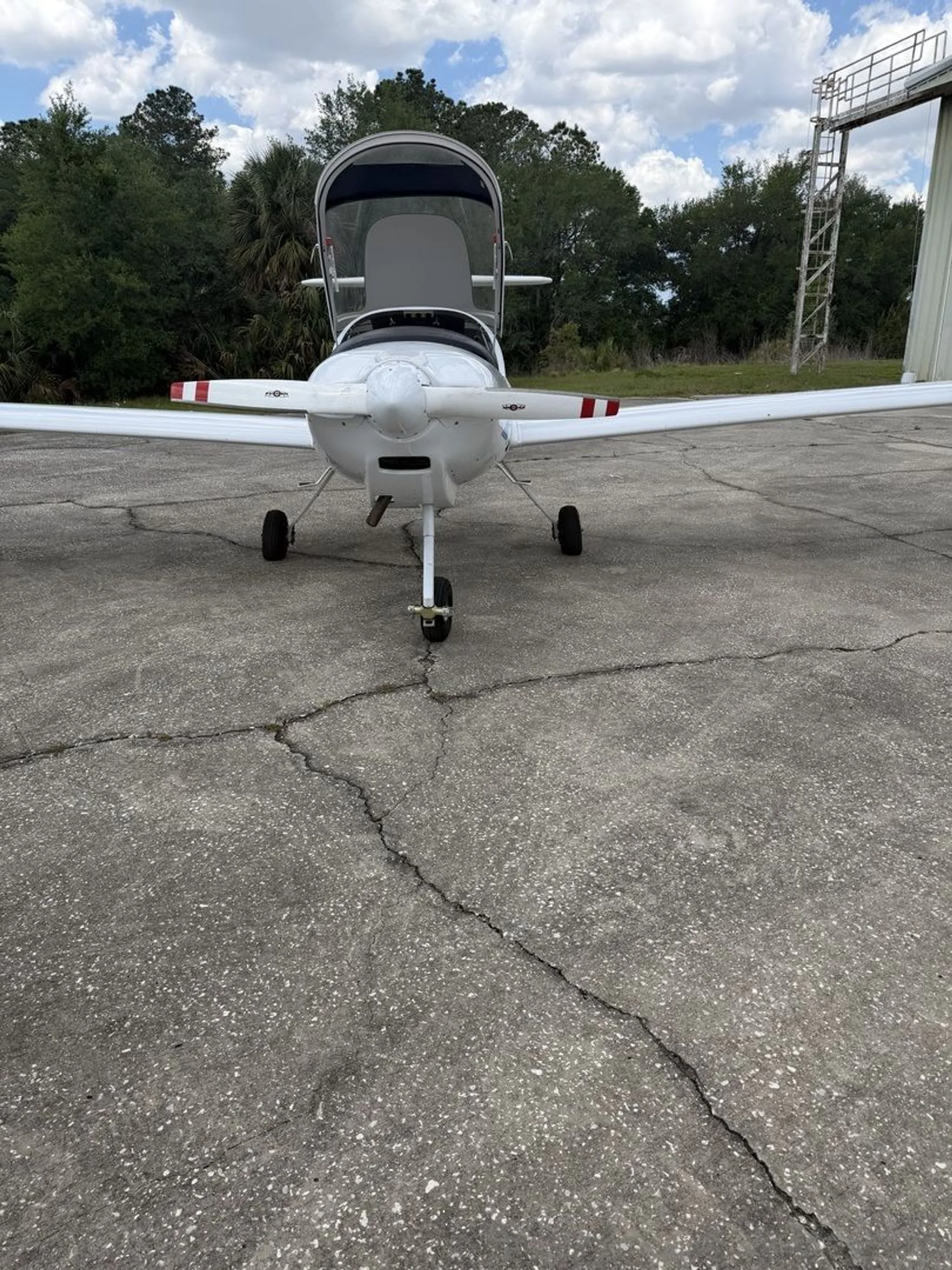 A white Diamond DA20-C1 Eclipse aircraft with red stripes on the wings, seen from the front with its canopy open on an asphalt tarmac.